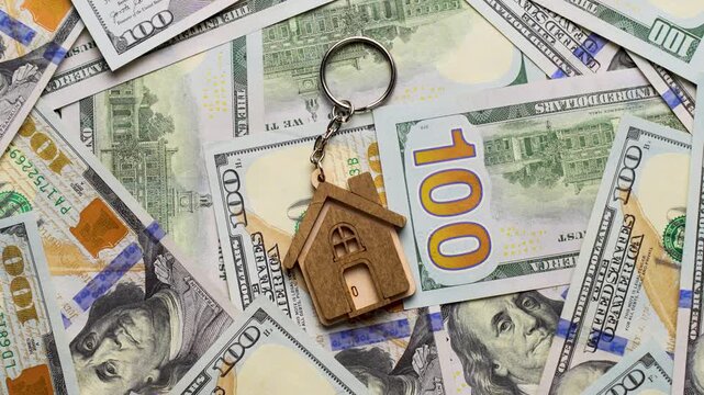 Property Investment and Home Ownership Concept. A wooden house shaped keychain with silver keys lying on a background of scattered dollar bills