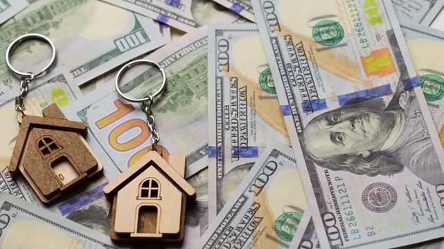 Property Investment and Home Ownership Concept. A wooden house shaped keychain with silver keys lying on a background of scattered dollar bills