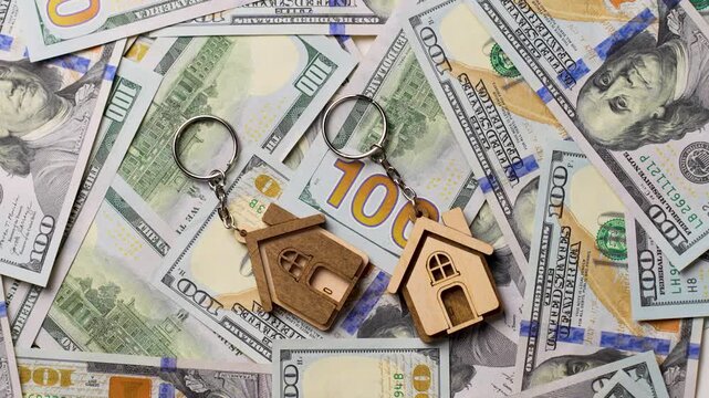 Property Investment and Home Ownership Concept. A wooden house shaped keychain with silver keys lying on a background of scattered dollar bills