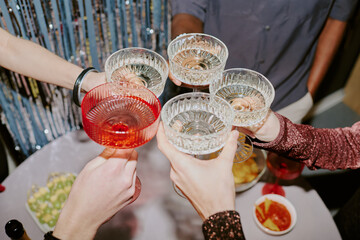 Naklejka premium Diverse group of young adult and middle aged hands toasting with crystal glasses at festive celebration, visible snacks and drinks on table, multiethnic gathering enjoying party