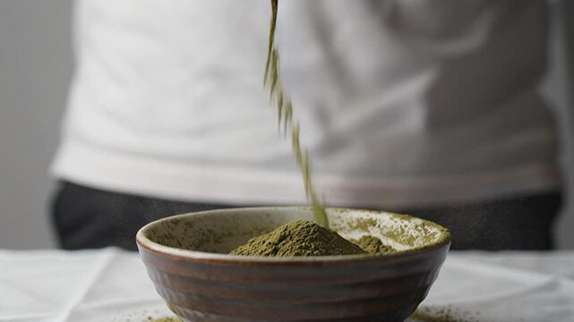 Person pouring vibrant green matcha tea powder from small glass jar into rustic ceramic bowl for traditional ritual prep.