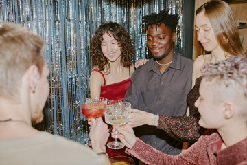 Naklejka premium Group of young adult multiethnic friends celebrating together, standing close and clinking cocktail glasses, smiling and enjoying festive moment at party with shimmering backdrop