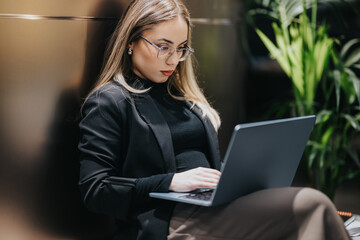 A professional woman uses a laptop, focused and composed in a contemporary office setting. She sits with calm concentration among greenery, conveying productivity and modern work style.