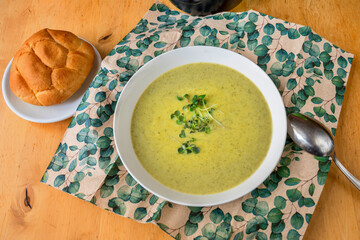 Creamy broccoli soup in white plate with spoon, napkin and bun.