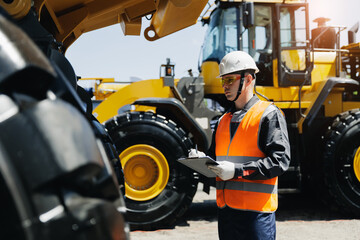 Young caucasian male construction worker in safety gear checks tread depth on tire of industry truck