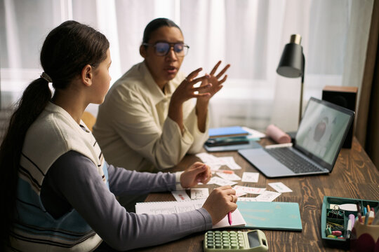 Teenage Caucasian girl sitting at desk studying with middle aged Black woman tutor explaining lesson using flashcards and laptop, both focused on educational activity in home setting