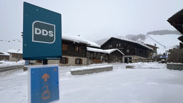 Livigno, Italy - February 4, 2026: Milano Cortina 2026 DDS sign and directional panel in snowy winter village with traditional wooden houses and ski slope in Livigno, Italy