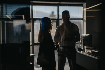 Business people exchange ideas over documents in a dim office. Business employees stand by the window, discussing work.