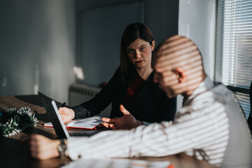 Two business people sit at a table with a laptop and papers, engaged in discussion. A daytime office scene shows one business person listening while the other explains a point.