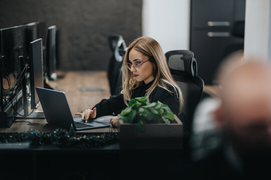 Business employee works on a laptop at a desk with multiple monitors. Another business person appears blurred in the foreground in a modern office.