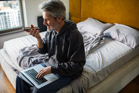 Middle aged man using laptop and voice assistant at home