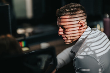 A business professional sits at a desk with striped light on his face. A business employee works in a dim office with shadows from blinds.