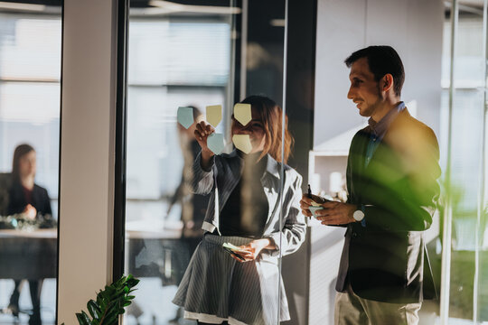 Two business people collaborate in a glass-walled office. They place sticky notes on the glass as they discuss ideas in a professional setting.