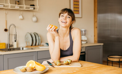 Woman in sportswear, enjoying a fresh orange in her kitchen, surrounded by healthy fruit