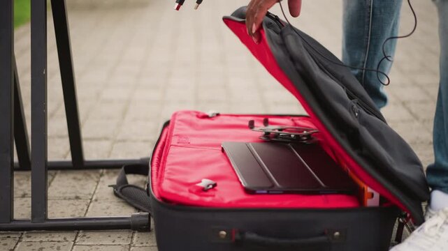street musician opens red suitcase gear hands plug controller into mixer jeans and white sneakers visible pavement backdrop urban square atmosphere casual preparation for live performance cables