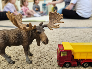 Obraz premium Toy truck is next to a toy moose. The truck is red and yellow. The moose is brown and has a large head. The scene takes place on a carpeted floor, with the children sitting in kindergarten
