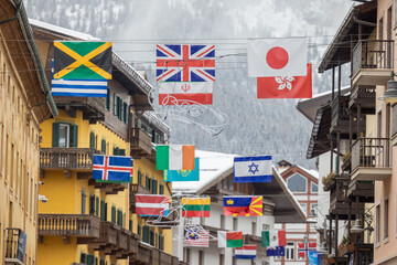 International flags hang above a snowy street in Cortina d Ampezzo, set against Alpine chalets and misty mountains, symbolizing global unity, winter tourism, and Olympic celebration.