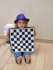 Child is holding a checkered chess piece of paper. The child is wearing a purple hat near globe