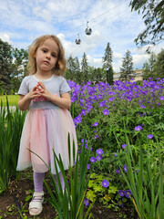 Little girl is standing in a garden with purple flowers. She is wearing a rainbow dress near cableway