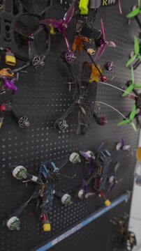 Diverse collection of custom built fpv racing drones and freestyle quadcopters hanging neatly on black pegboard wall in a workshop, showcasing various frames, propellers, and drones components