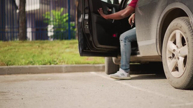 black man in red shirt stepping out of dusty car in sunlit parking lot, aspiring DJ in jeans and sneakers scans street with thoughtful expression, cinematic closeup shows urban fence and quiet
