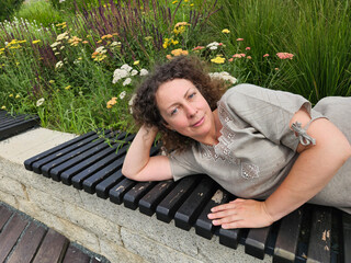 Woman is laying on a bench in a park. She is wearing a tan dress and has her curly hair in a ponytail