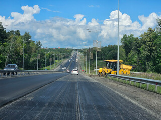 Yellow road asphalt spreader is on the road. There are many cars on the road with removed top layer of asphalt