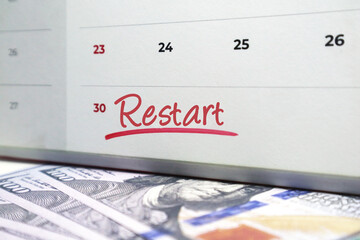 White spiral calendar with the word Restart handwritten in red ink, lying on a background of scattered US one hundred dollar currency notes.