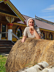 Woman is sitting on a pile of hay. She is wearing a white hat. The hay is brown and is piled up in front of a building with platbands and wood-carved cornices