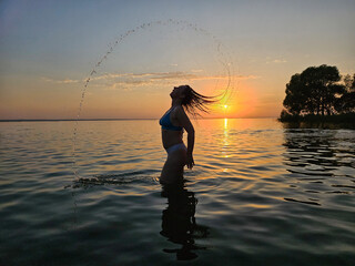 Woman is in the water, splashing water. The sun is setting in the background.