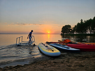 Man boy with tube cart is walking on the sunset beach with a surfboard. There are three surfboards on the beach, one of which is yellow