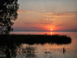 Lake with a sunset in the background. The sun is setting and the sky is orange. The water is calm and there are trees in the background