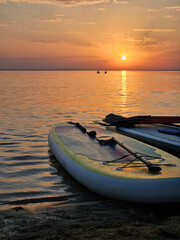 White surfboard is laying on the sand next to the water. The sun is setting and two silhouette people on supboards