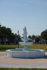 Elegant White Sculpture Fountain in a Park Setting with Water Droplets and Ornate Tiered Design. Pristine white, multi-tier fountain stands in a sunlit park, water gently spills over the edges creatin