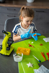 Girl wearing protective gloves examining grown crystal at table with microscope and laboratory...