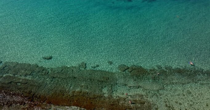 Aerial view of a sandy beach featuring neatly arranged rows of beach umbrellas. The shoreline reveals clear turquoise water with visible rocky formations under the water surface. Summer morning.