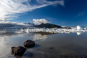 Bright sunlight glows across Jökulsárlón glacier lake as blue and white icebergs drift beneath Southern Iceland’s towering mountains and soft clouds mirrored in the still midday water © lightcaptured