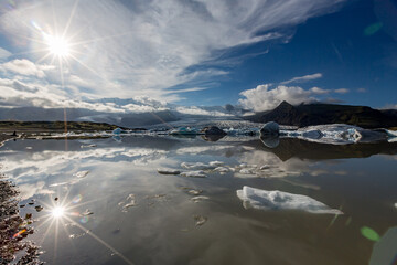 Bright sunlight glows across Jökulsárlón glacier lake as blue and white icebergs drift beneath Southern Iceland’s towering mountains and soft clouds mirrored in the still midday water © lightcaptured