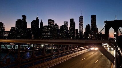Brooklyn bridge roadway at night with light trails from moving vehicles and a illuminated lower manhattan skyline featuring skyscrapers and one world trade center under a fading dusk sky © DawDunia