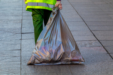 Worker in high-visibility uniform carries a bulky garbage bag across a tiled city pavement. The scene captures everyday urban maintenance, highlighting the often-overlooked effort behind keeping publi