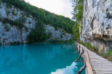 Obraz premium Plitvice Lakes, Croatia - August 16, 2025: Tourists walking on a wooden boardwalk over turquoise water. Scenic landscape with limestone cliffs and a cave in the National Park