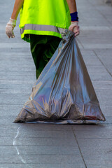 City worker in a bright reflective uniform and protective gloves is seen carrying a large, filled garbage bag across a paved street.