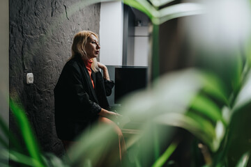 A professional woman sits in a modern office, talking softly. The scene shows her near a computer with lush plants framing the foreground.