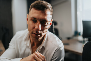 A focused man in a white shirt leans forward, holding a pen at a desk in a contemporary office. The scene captures a thoughtful expression with a blurred workstation in the background.