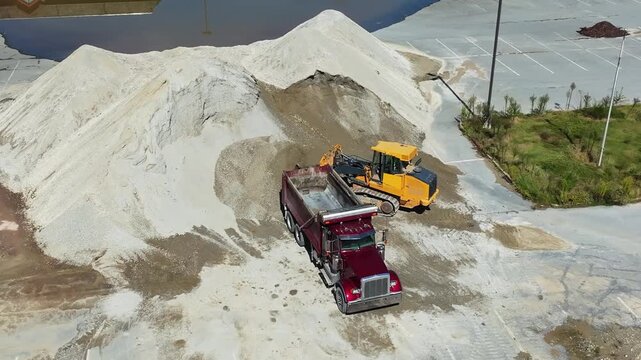 Trucks and bulldozers work at a construction site on a summer afternoon. They transport and move sand piles near a pond. Machines operate efficiently on the site.