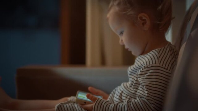Young child sits snug on sofa, absorbed vibrant cartoons on smartphone screen during peaceful evening, raising concerns about potential harm to their developing eyesight from prolonged screen exposure