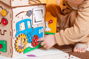A small child plays with a colorful wooden toy house. The child has blond hair and is wearing a beige suit. The busy board toy has gears and various shapes.