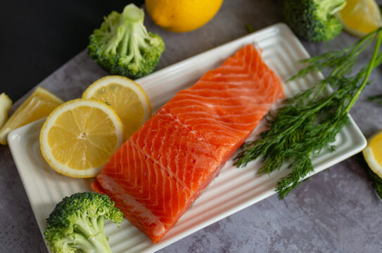 Raw trout fillet plated on a white ceramic dish with lemon slices (citrus limon), fresh dill (anethum graveolens), and broccoli (brassica oleracea). Bright food styling for seafood dinner ideas, nutri