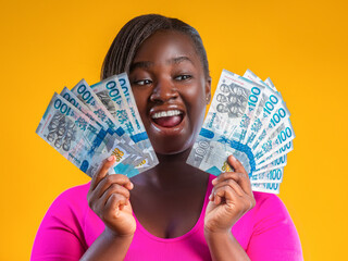 A joyful Black woman holds fans of Ghana cedi currency notes against a vibrant yellow background. She wears a pink top, her expression beaming with excitement and success as she celebrates her wealth