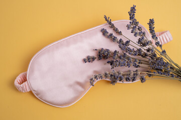 Pink silk eye mask with dried lavender flowers on yellow background. Natural sleep aid and...